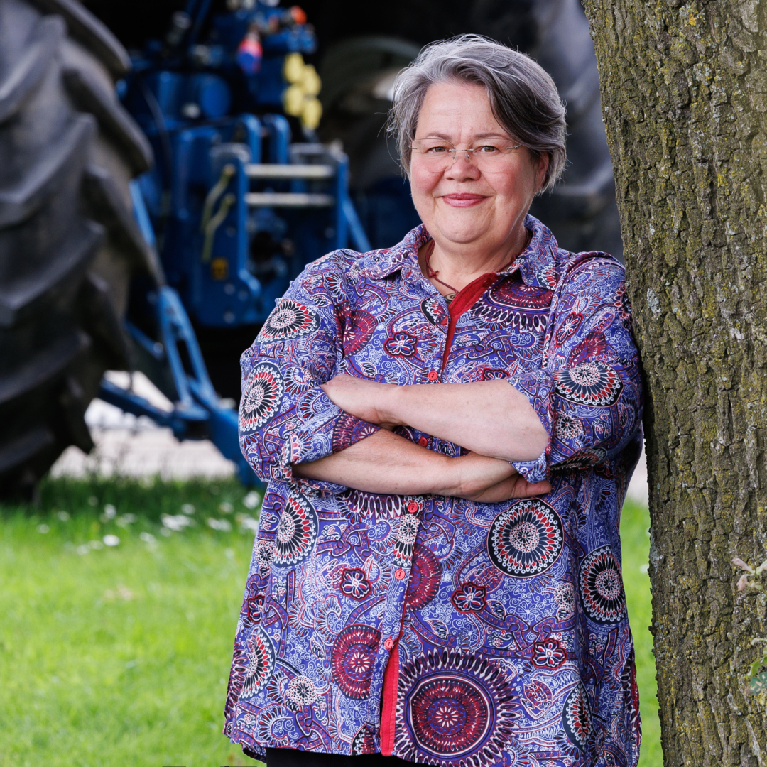 Ansjeliek Coolbergen, leunend tegen een boom met een vriendelijke glimlach. Ze draagt een kleurrijke blouse met een opvallend patroon en staat buiten in een groene omgeving met een tractor op de achtergrond.