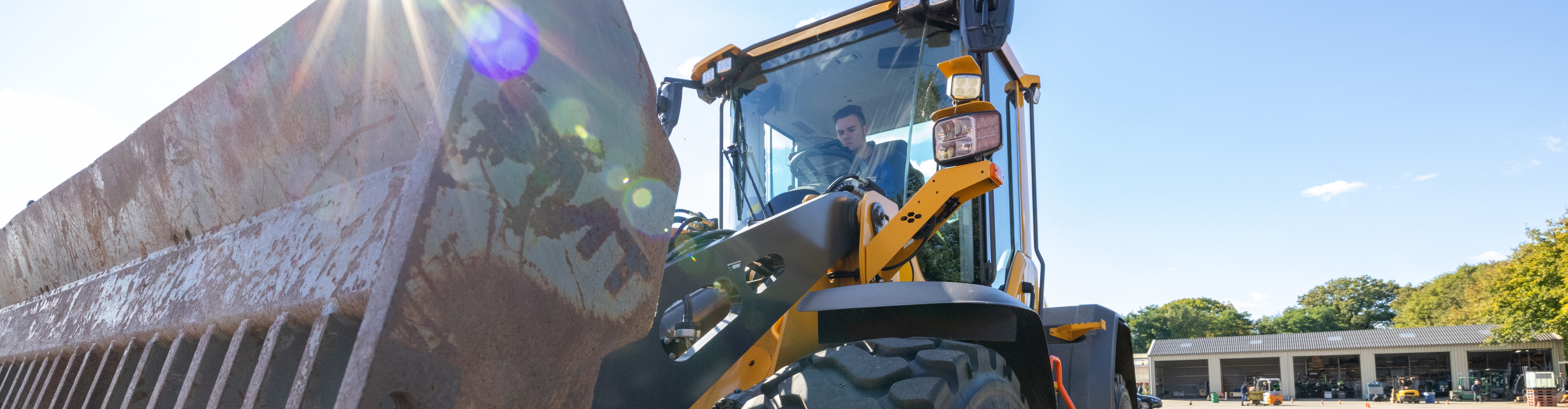 Een gele Volvo-shovel met het logo van SMT staat op het terrein van Aeres Tech onder een heldere blauwe lucht. De zon straalt over de grote metalen bak van de machine. In de cabine bestuurt student Tosh Frijns de shovel.