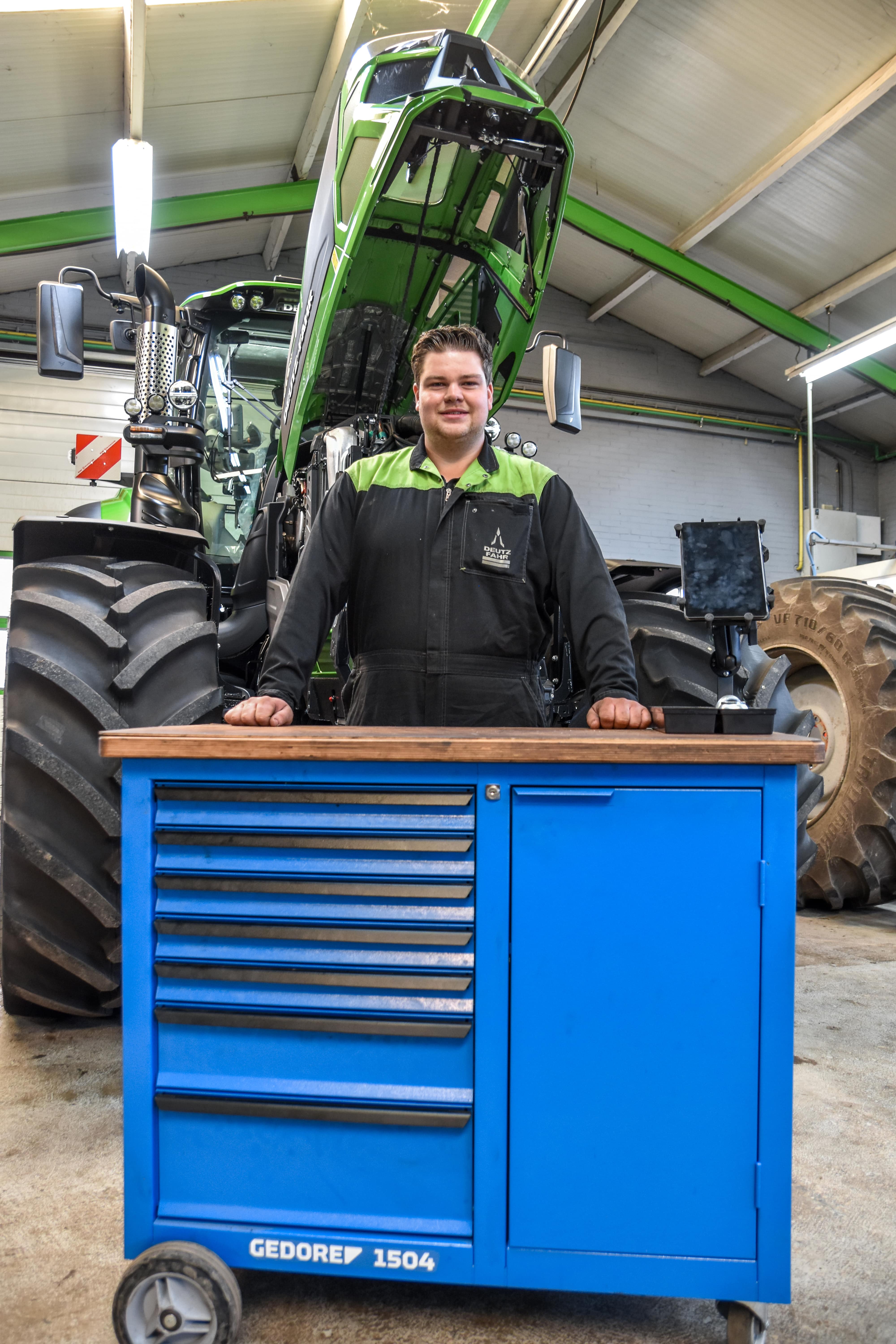Nick van der Slot staat in een werkplaats naast een opengeklapte groene tractor, met op de voorgrond een blauwe gereedschapskist. De werkplaats heeft een industriële uitstraling met veel verlichting.