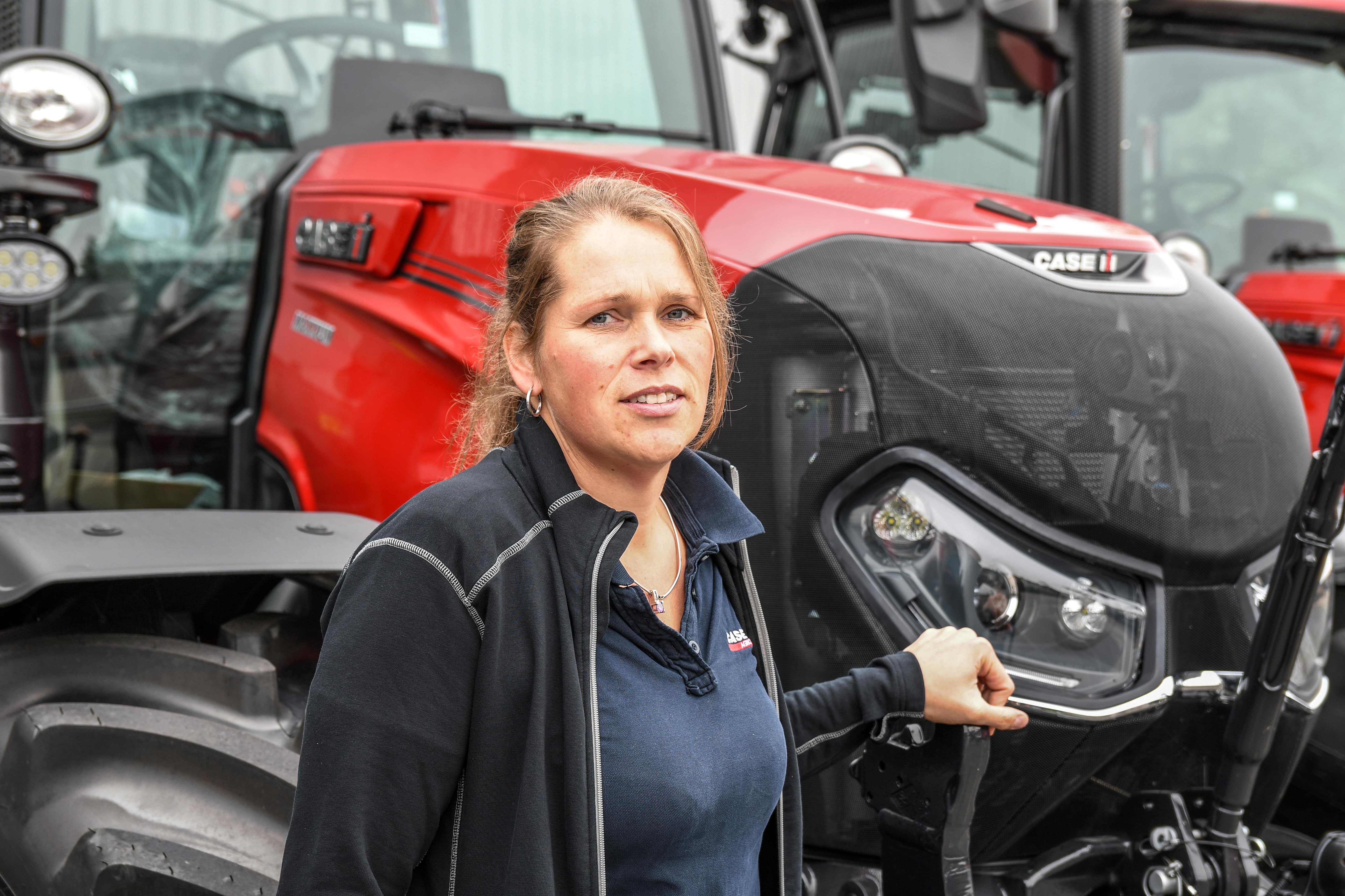Lisette Vos staat in werkkleding naast een rode Case IH-tractor en kijkt in de camera. Op de achtergrond zijn meer tractors zichtbaar.