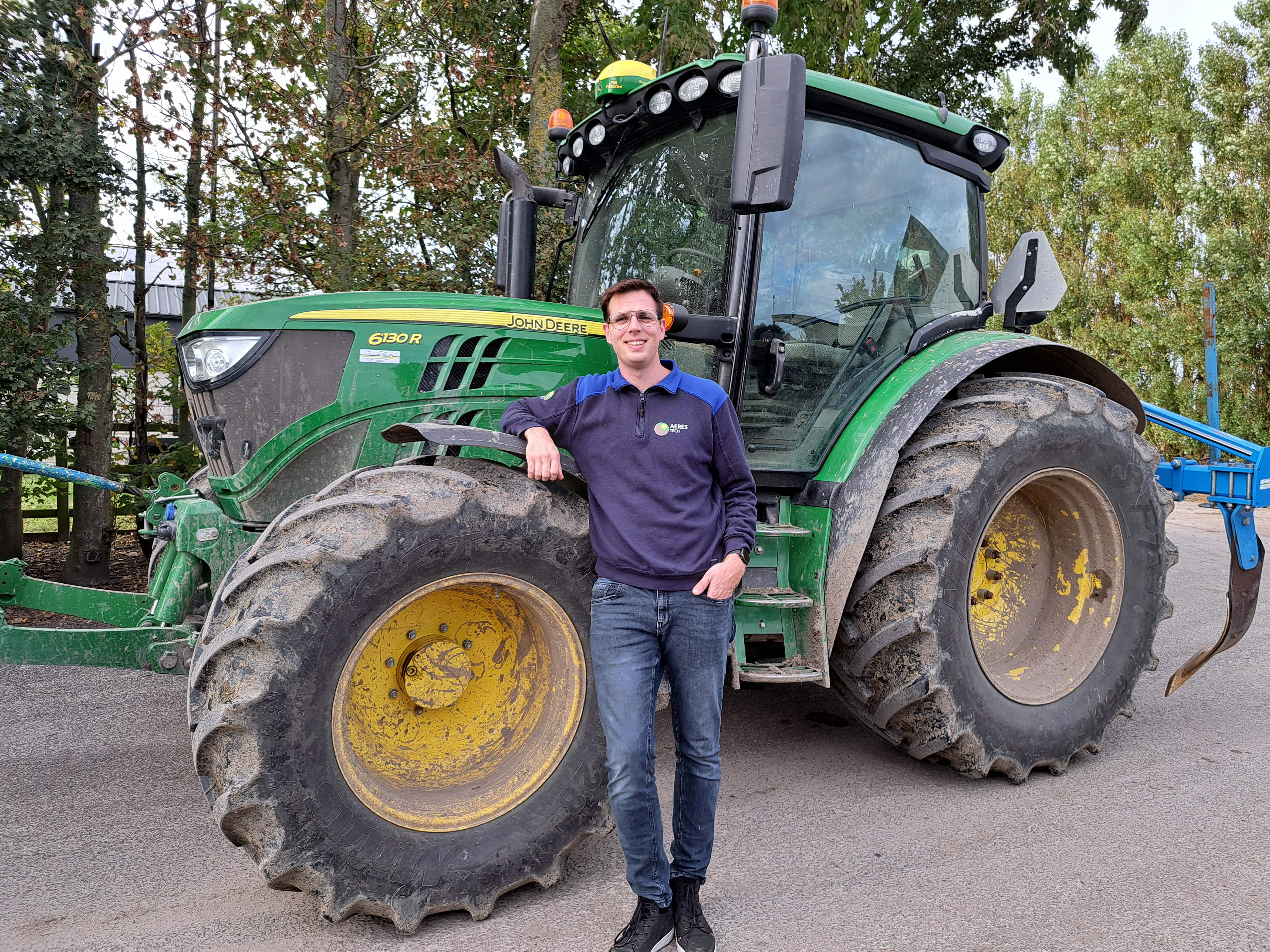 Stef van Gerven staat tegen een groene John Deere-tractor in een blauwe trui met het Aeres Tech logo erop, met bomen en een boerenerf op de achtergrond.