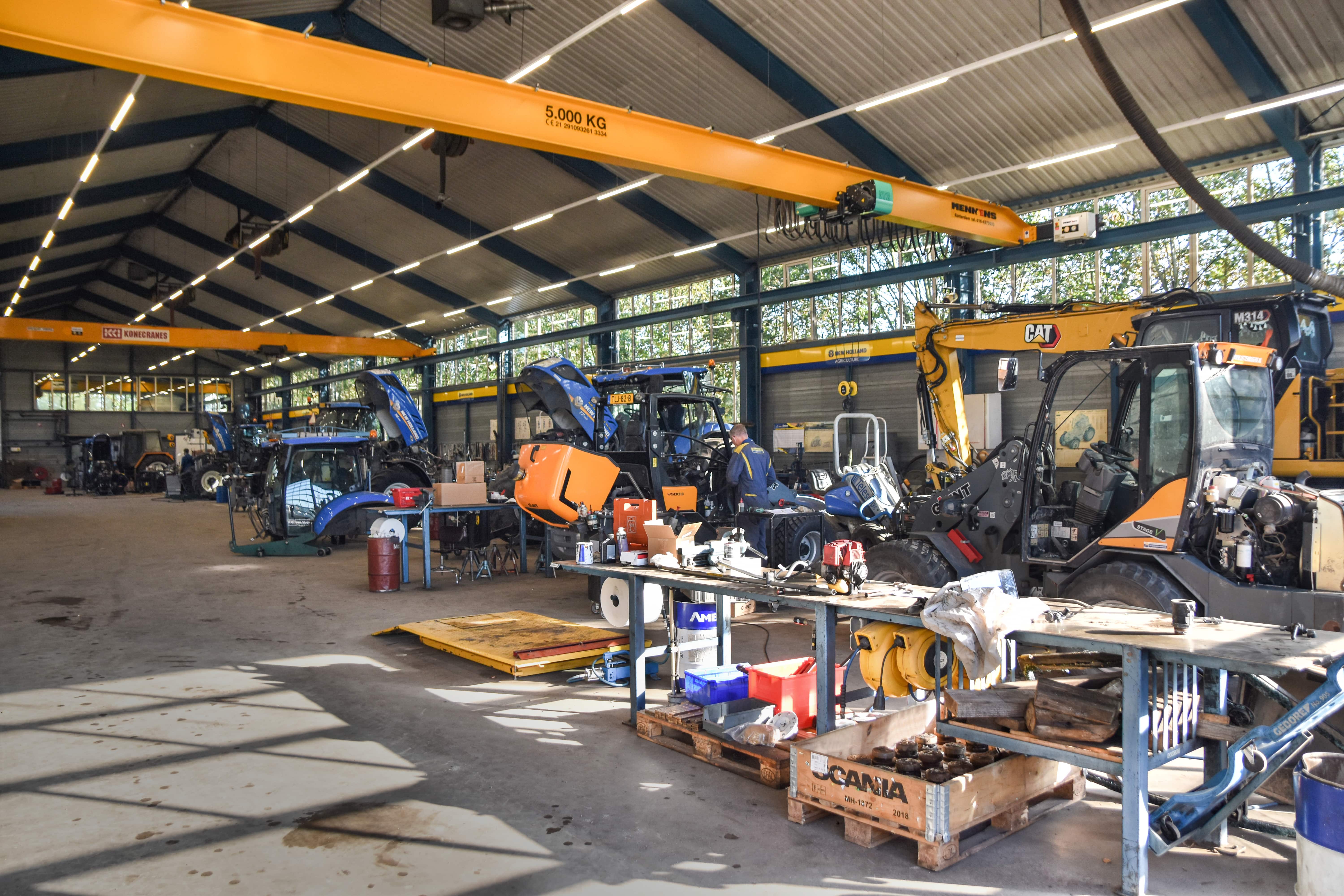 Een grote, goed verlichte werkplaats van Uittenbogerd Heukelum met verschillende landbouwmachines en bouwmachines. Monteurs zijn aan het werk tussen de gereedschappen en werkbanken, onder een gele kraan met een capaciteit van 5000 kg.